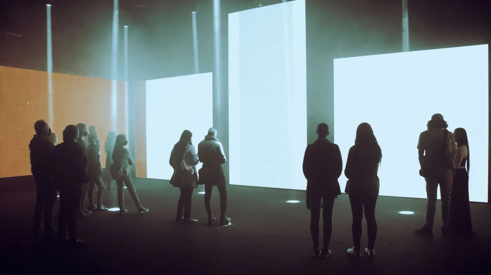 Visitors in silhouette stand before large illuminated screens in a dark exhibition hall.